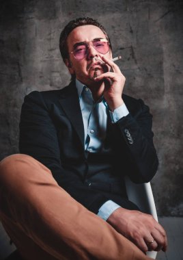 Resmi takım elbiseli ve füme mavi gözlüklü vahşi bir adam oturmuş sigara içiyor.
