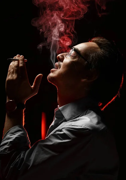 Жестокий небритый мужчина в официальной рубашке и очках наслаждается курением и выдуванием дыма изо рта — стоковое фото