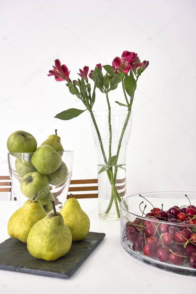 fresh apples and pears near to vase of cherries on the table in the kitchen together to vase of red fresh flowers