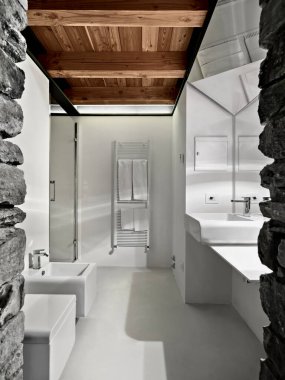 Modern banyo, taş duvarlar ve reçine zemini sağda tezgah lavabosu solda bide ve arka planda tuvalet radyatör ve duş kutusu tavanda ahşap kirişler var.