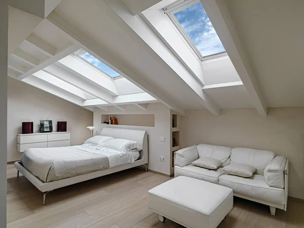 手前の近代的な屋根裏部屋の寝室の内部ショット背景にフットレスト付きの革のソファがあり2つの天窓によって照らされたベッドがあります — ストック写真