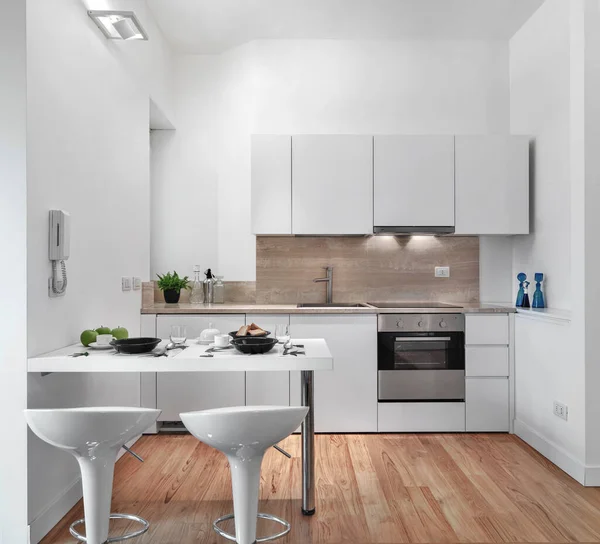 modern total white kitchen interior, with wooden floor in the foreground two stools and a dining table, in the background the kitchen wall units on the wall, and the sink cabinet plus the built-in oven