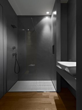 Arka planda ahşap zemin olan modern banyo. Sağda reçine duvarları olan cam duş bölmesi. Tezgahın üstünde lavabo.