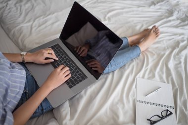 Kadın yatakta dizüstü bilgisayar kullanarak. Genç Asyalı kız yatak kağıt Not defteri, kalem ve yatakta gözlük ile dizüstü bilgisayar üzerinde çalışan.