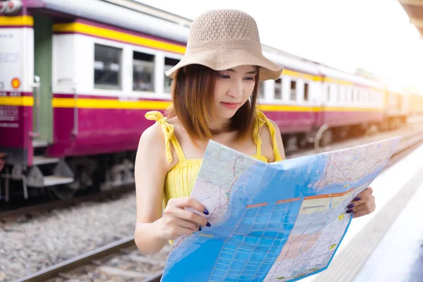 Asian traveler woman looking map at train station find destination. Happy backpacker girl lost in the city search on map. Hipster photographer lady waiting for train on vacation. People using rail road transportation on holiday.