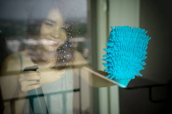 Young woman washing window. happy maid in gloves holding bottle spray cleaning window with blue rag duster and cleanser detergent at home. People housework and housekeeping glass cleanup concept.