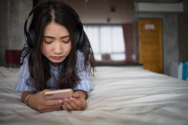 Genç kız evde yatakta smartphone üzerinden müzik dinleme. Asyalı kadın yatakta metin mesajı telefon ederken yatak odasında cep telefonu kulaklık ile müzik dinleme holding yalan.