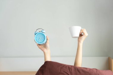 Kol Yatak odası, Genç kız iki elle battaniye yapışkanlık dışarı kahve fincanı ve yorgan arkasında mavi çalar saat dayanabiliyor kaldırdı gösterilen kadın. sabah kavram eğlenceli uyandırmak.