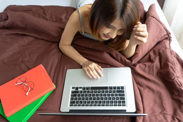 Happy woman lying down on bed with laptop and books.Top view mockup woman laying on bed using and typing on laptop keyboard.Young Asian girl relaxing in bed on lazy day. Lifestyle education concept.