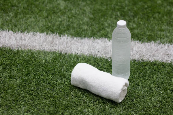 Water bottle sport drink and towel in football field on green artificial grass background.