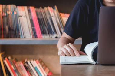 Genç iş adamı hisse senedi ticaretini öğreniyor. Adam kütüphanede oturmuş, parmak işareti yapıyor, kitap okuyor ve dizüstü bilgisayar kullanarak internete bağlanıyor..