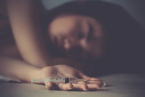 アジアの女の子の薬物中毒者は、落ち込んで絶望的な汚い部屋の床に横たわっています。バックグラウンドで中毒若い女性の薬物で床に用量注射を行う注射器を保持する意識不明の女性の手をクローズアップ. — ストック写真