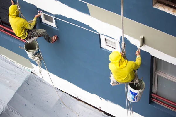 Pintores colgando en rollo, pintando color en la pared del edificio. constructor de fachada trabajador con cepillo de rodillos, trabajando en el edificio alto. construcción de seguridad con correa de elevación en la ciudad . — Foto de Stock