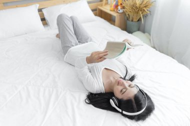 Yatakta kulaklıkla dinlenen genç bir kadın. Asyalı kız yatakta uzanıp müzik dinliyor ve kitap okuyor. Bayan favori kitaptan hoşlanıyor, rahat bir yatakta dinleniyor, rahatlıyor ve hayat tarzı kavramını kullanıyor..