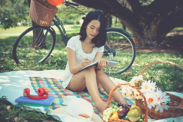 Young woman picnic in park with fruit basket writing note book under tree. Asian girl with flower, book ,coffee cup and headphones. Happy female in garden with bicycle in background. lifestyle in green nature outdoor.