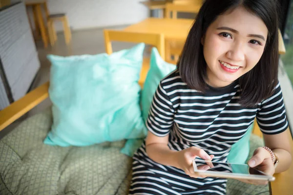 Młoda kobieta relaks za pomocą tabletu uśmiech patrząc na kamerę. Happy Asian Girl siedzi na kanapie trzymając tablet komputera w ręku. Ludzie styl życia z koncepcją technologii. — Zdjęcie stockowe