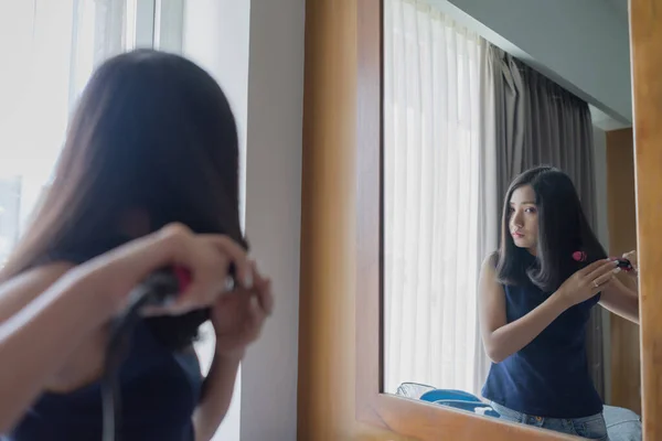 Young Asian woman straightening hair with hair straightener flat iron while looking into the mirror at home.