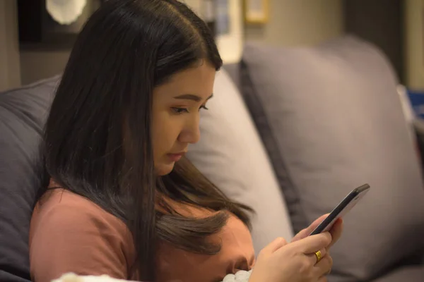 Giovane donna felice utilizzando smartphone seduto sul divano a casa. asiatico utilizzando app ragazza sms su cellulare . — Foto Stock