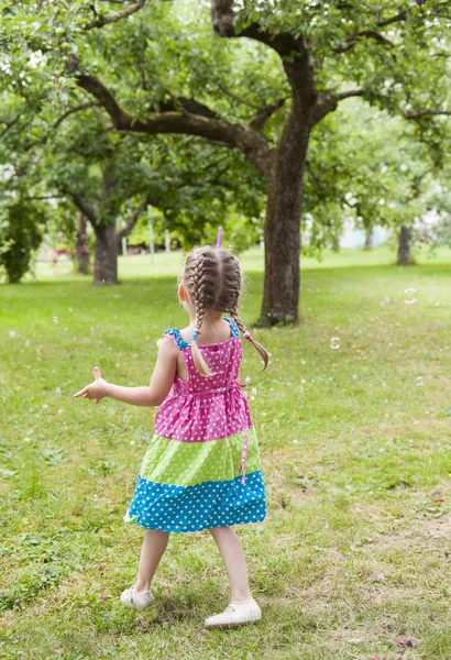 シャボン玉と公園で踊るカラフルなドレスでおさげの少女 — ストック写真