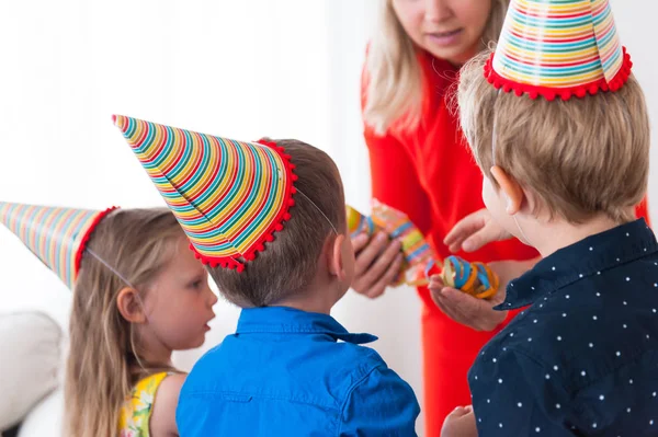 Two boys and one girl in party hats turned away from the camera take bright confetti from moms hand