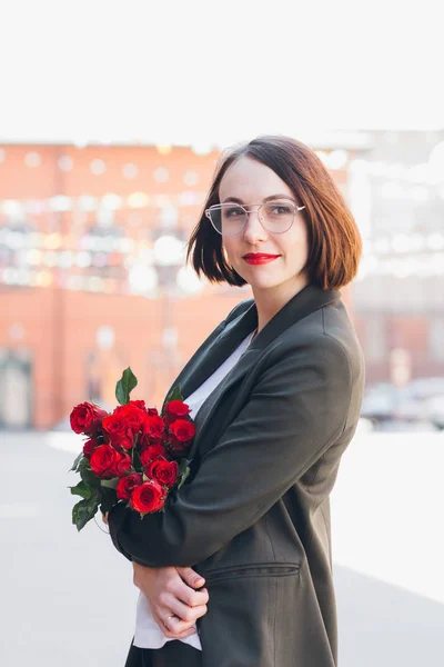 Young beautiful woman in a business suit and with a bouquet of red roses posing on the streets. Toning.