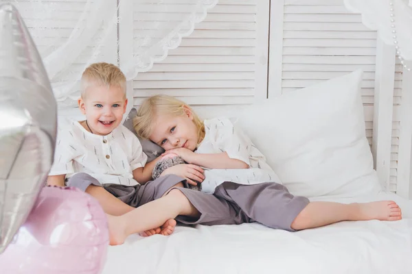 Little sister and brother sit on the bed in outfits. Holiday with balloons. Toning.