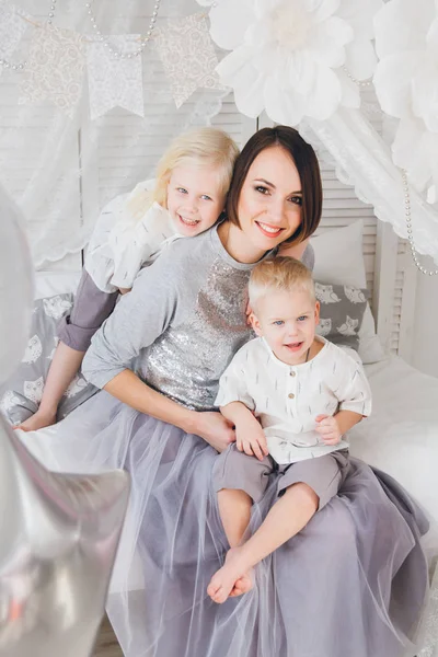 Mutlu anne, kızı ve oğlu yatakta kıyafetler içinde oturuyor. Balonlar ile tatil. Tonlama.