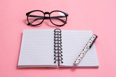 Not defteri, kalem ve pembe bir arka plan üzerinde gözlük. Düz ley kompozisyon. Masanın üzerinde ofis, banka donatımı.