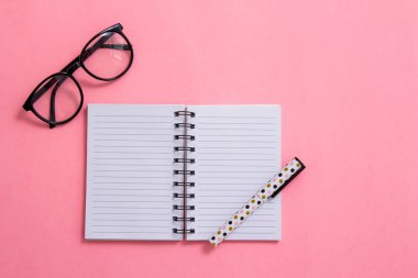 Not defteri, kalem ve pembe bir arka plan üzerinde gözlük. Düz ley kompozisyon. Masanın üzerinde ofis, banka donatımı.