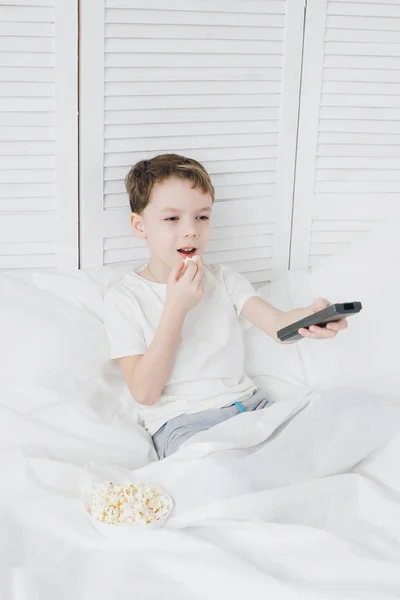 Мальчик ест попкорн, сидит в постели и смотрит телевизор — стоковое фото