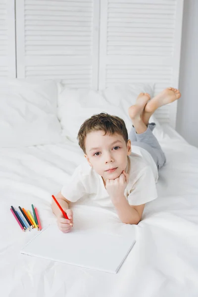Мальчик рисует цветными карандашами, сидя на кровати — стоковое фото