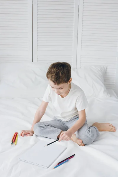 Мальчик рисует цветными карандашами, сидя на кровати — стоковое фото