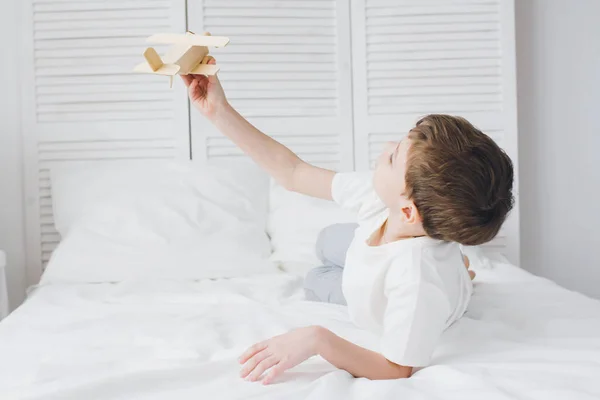 Симпатичный мальчик играет с деревянным самолетом, сидя на кровати — стоковое фото