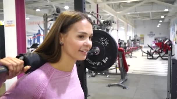 Vrolijk jong meisje hurkt met een lange halter in de sportschool — Stockvideo