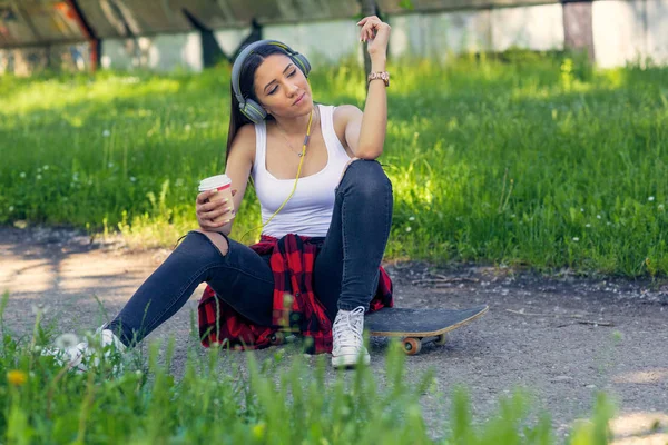 Sporty skateboarder girl sitting on skateboard drink coffee  and listing music