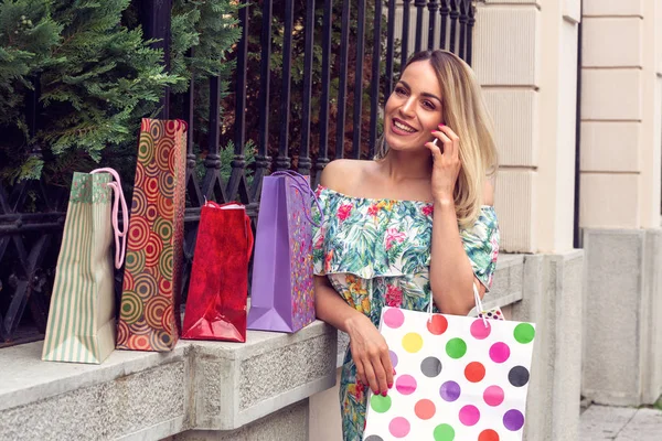 Kobieta trzyma torby na zakupy, za pomocą inteligentnego telefonu i uśmiechając się stojąc na zewnątrz — Zdjęcie stockowe