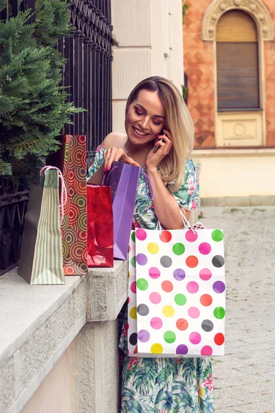 Uśmiechnięta kobieta trzyma torby na zakupy, za pomocą inteligentnego telefonu i uśmiechając się stojąc na zewnątrz — Zdjęcie stockowe