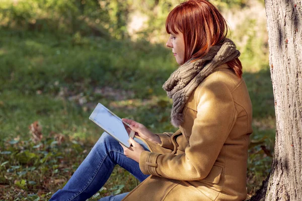 Mujer sentada bajo un árbol en el parque de otoño con la tableta — Foto de Stock