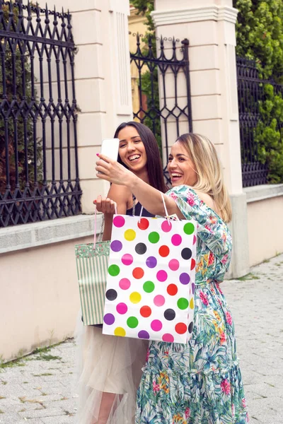 Chicas sosteniendo bolsas de compras, usando un teléfono inteligente y sonriendo mientras están de pie al aire libre — Foto de Stock