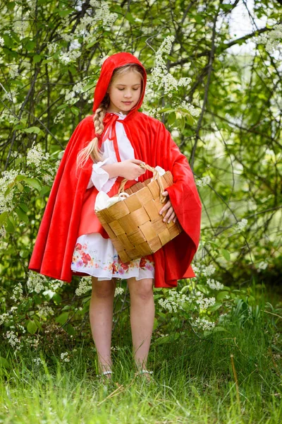 Lächelndes blondes Mädchen posiert in einem Kleid aus Rotkäppchen in der Nähe grüner Bäume — Stockfoto