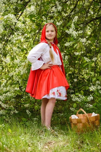 微笑的金发女孩摆在小红帽的礼服附近的绿树 — 图库照片