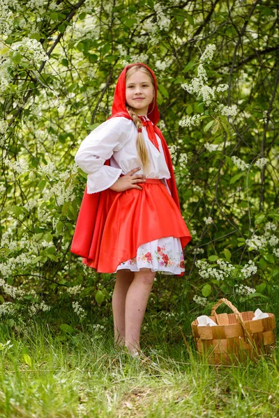 Lächelndes blondes Mädchen posiert in einem Kleid aus Rotkäppchen in der Nähe grüner Bäume — Stockfoto