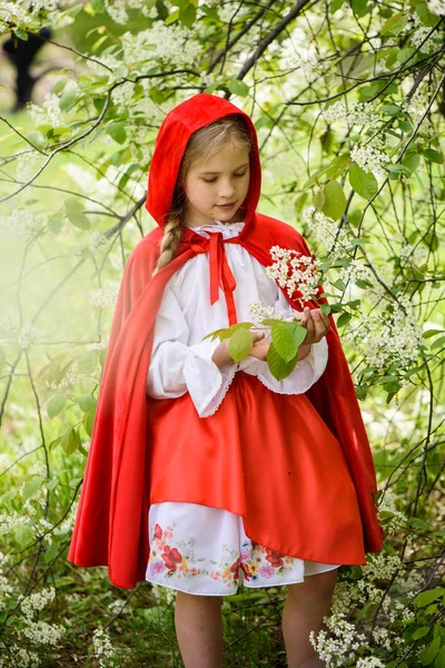 Lächelndes blondes Mädchen posiert in einem Kleid aus Rotkäppchen in der Nähe grüner Bäume — Stockfoto
