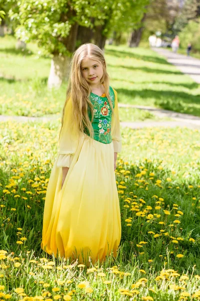 Blond meisje poseren in een geel groene jurk staande op het gras met paardebloemen gele bloemen — Stockfoto