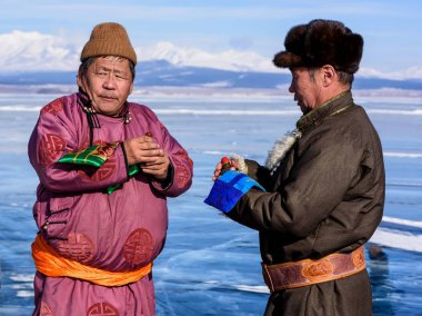 Hatgal, Mongolia, Febrary 23, 2018: mongolian people dressed in traditional clothing on a frozen lake Khuvsgul and change tobacco clipart