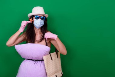 Şapkalı, maskeli, lastik eldivenli ve şapkalı bir kız Coronavirus izolasyonu sırasında yeşil arka planda deniz yıldızı ve plaj çantası tutuyor. Meydan okuma yastık elbisesi. Metin için boş alan. Komik yaz 2020