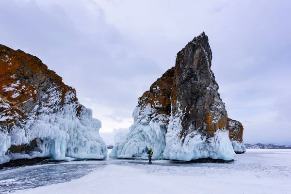 Lake Baikal, Rusia - 9 de marzo de 2020: La silueta de un hombre con chaqueta amarilla y mochila se encuentra cerca de dos rocas cubiertas de carámbanos. Lago Baikal en tiempo nublado . — Foto de Stock