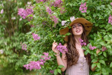 Pembe elbiseli, saman şapkalı kız yaz mevsiminin tadını çıkarıyor çiçek açan leylağın arka planında.
