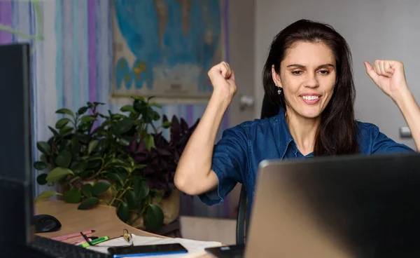 Overjoyed young woman in denim shirt working home with a laptop and stretching her arms up because got great news or won success. World map in the background