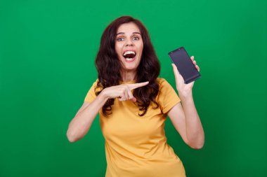 Genç ve çekici bir kadın elindeki boş telefon ekranını işaret ediyor ve kameraya bakıyor.